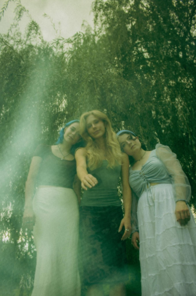 Three young women pose standing with heads together, wearing long skirts; a willow tree behind them