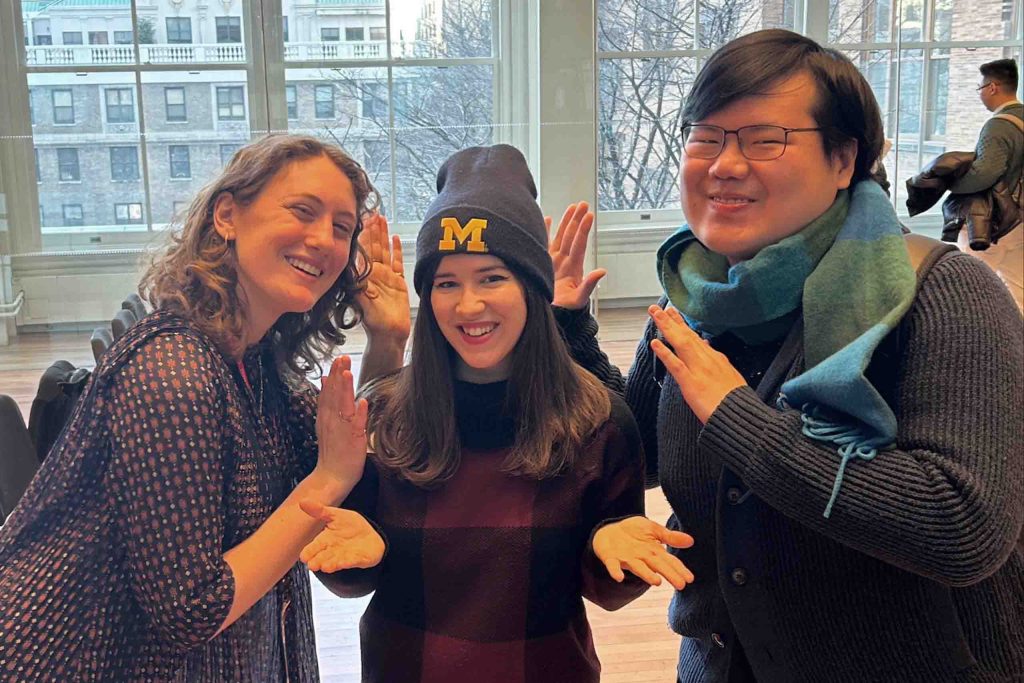 Three people pose and gesture to the Michigan hat in the center, in a city studio space