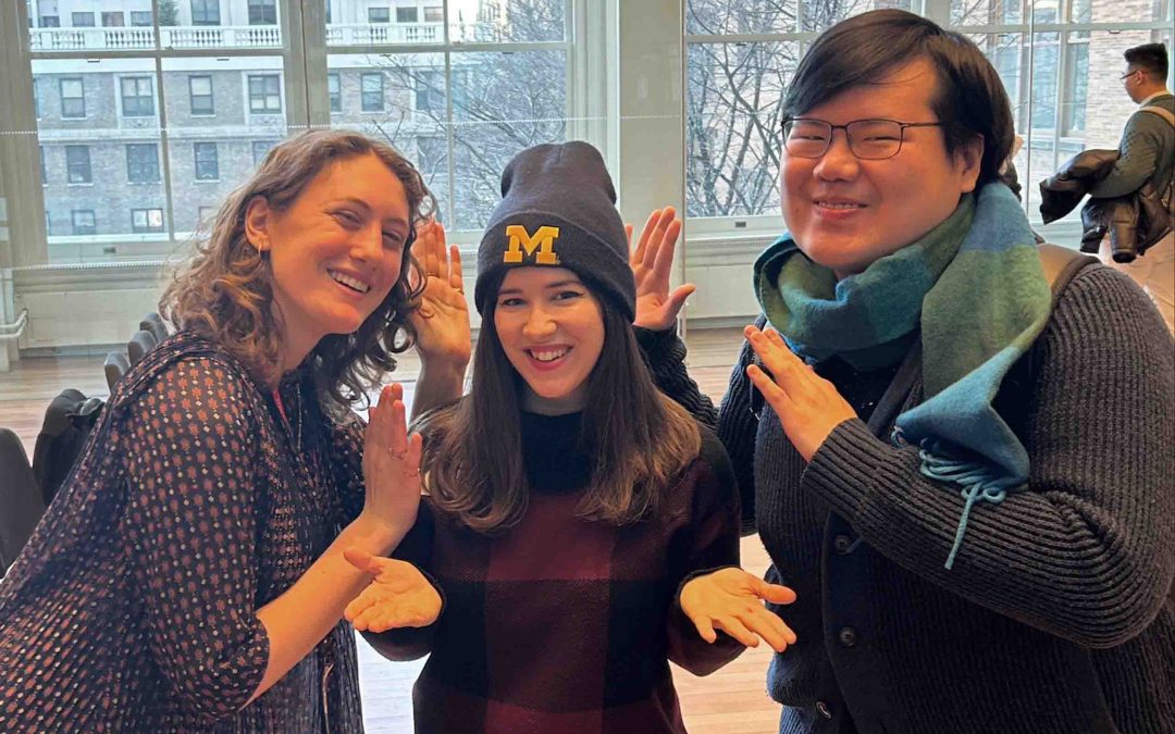 Three people pose and gesture to the Michigan hat in the center, in a city studio space