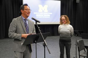 President Ono speaks from a stand and microphone as Prof. Shankel stands near him, with a U-M logo projection in background