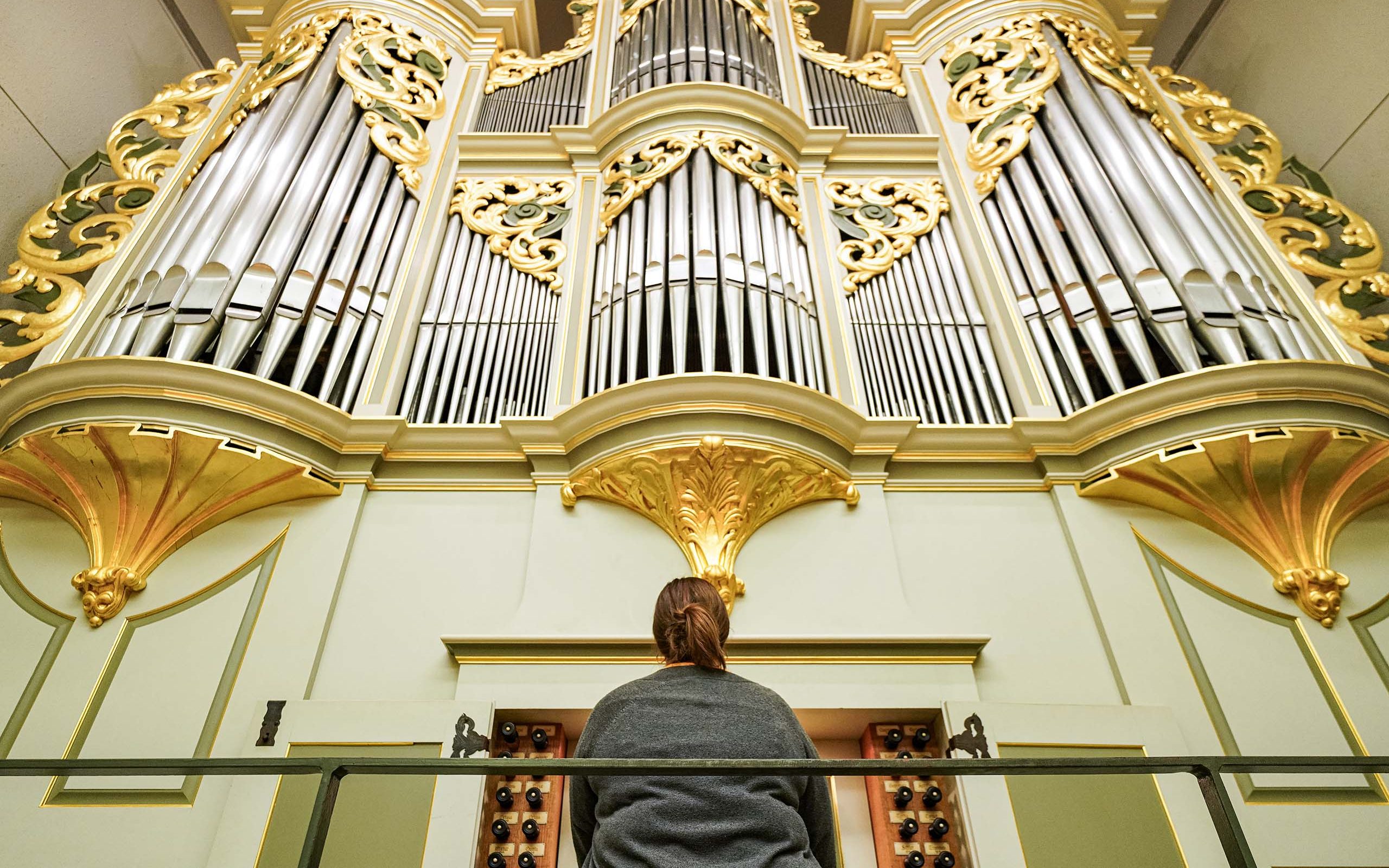 Organ player seated while playing, with silver organ pipes adorned with fancy gold trim rising high above them