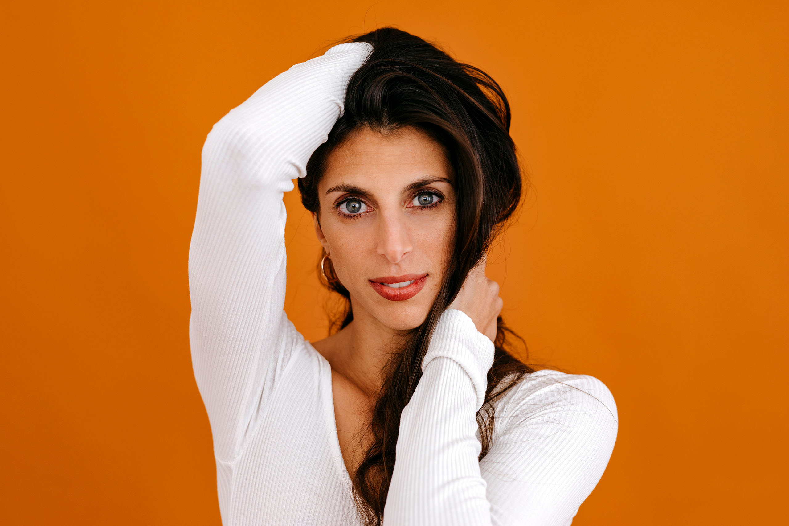 Catherine Coury studio headshot with arms folded up to her head and neck; wearing a white long sleeved shirt, with orange background