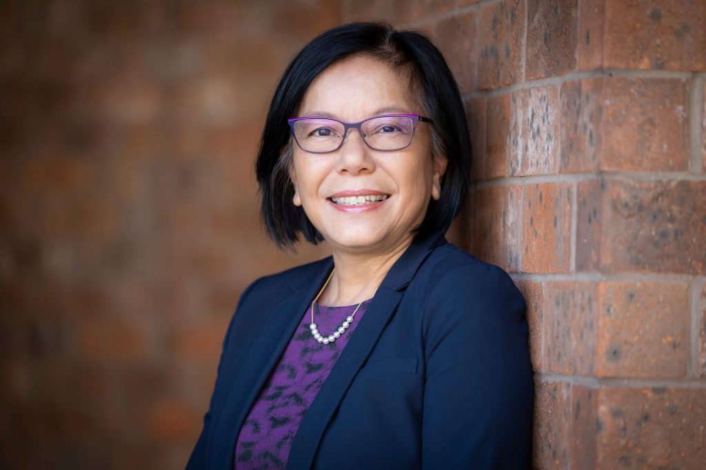 Nancy Rao headshot wearing suitcoat and purple shirt, brick wall in background