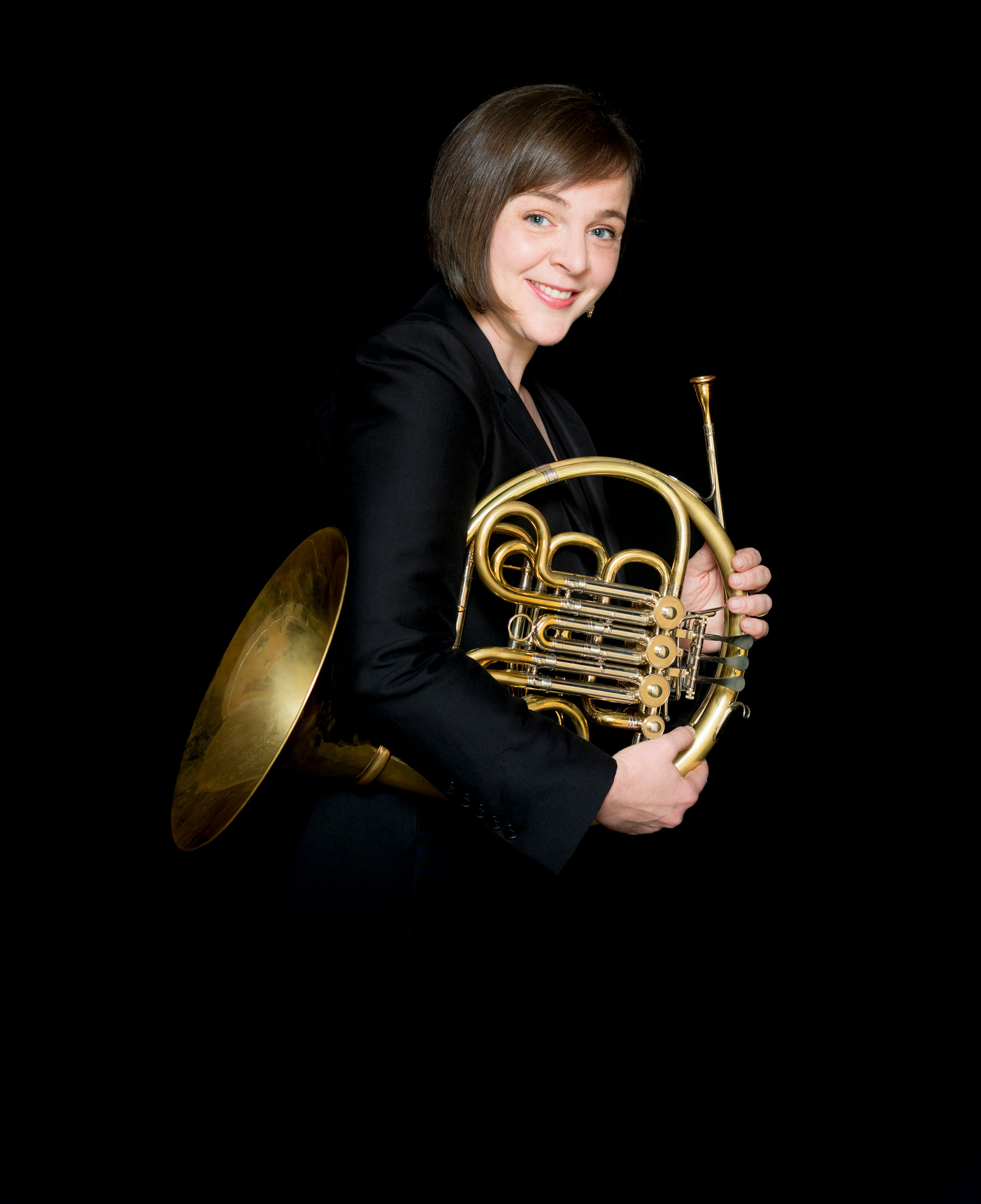 Rachel Childers studio portrait holding her French horn, wearing black with dramatic black background