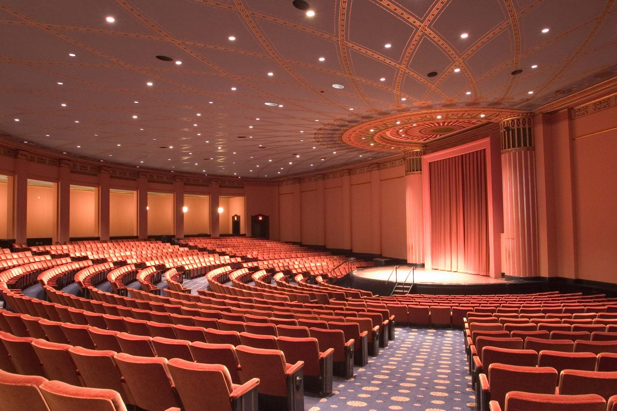 A semi-circular stage is surrounded by slightly sloped isles and plush theater seating