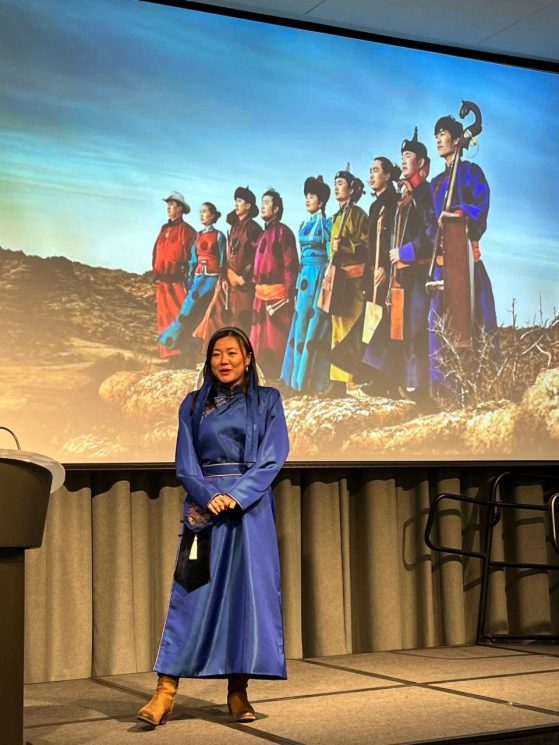 Sangseraima Ujeed speaks in front of a film projection, dressed in traditional Mongolian attire 