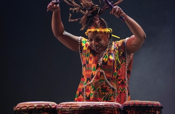 T. Ayo Alston performs drums on stage energetically, with arms raised holding percussion mallets