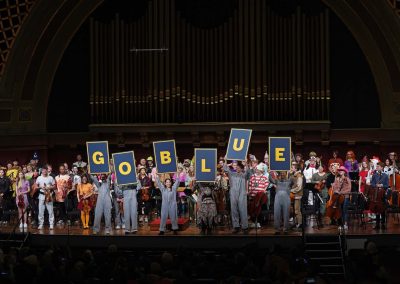 Orchestra on stage takes a bow wearing Halloween costumes; six people hold up large letters spelling GO BLUE