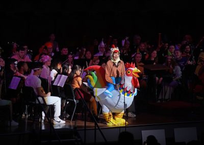 Graduate student conductor speaks to the audience wearing an inflatable chicken costume