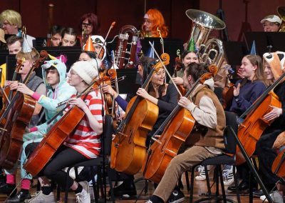 Smiling faces in the cello section on stage, including costumes of a unicorn, Where's Waldo, and a cat