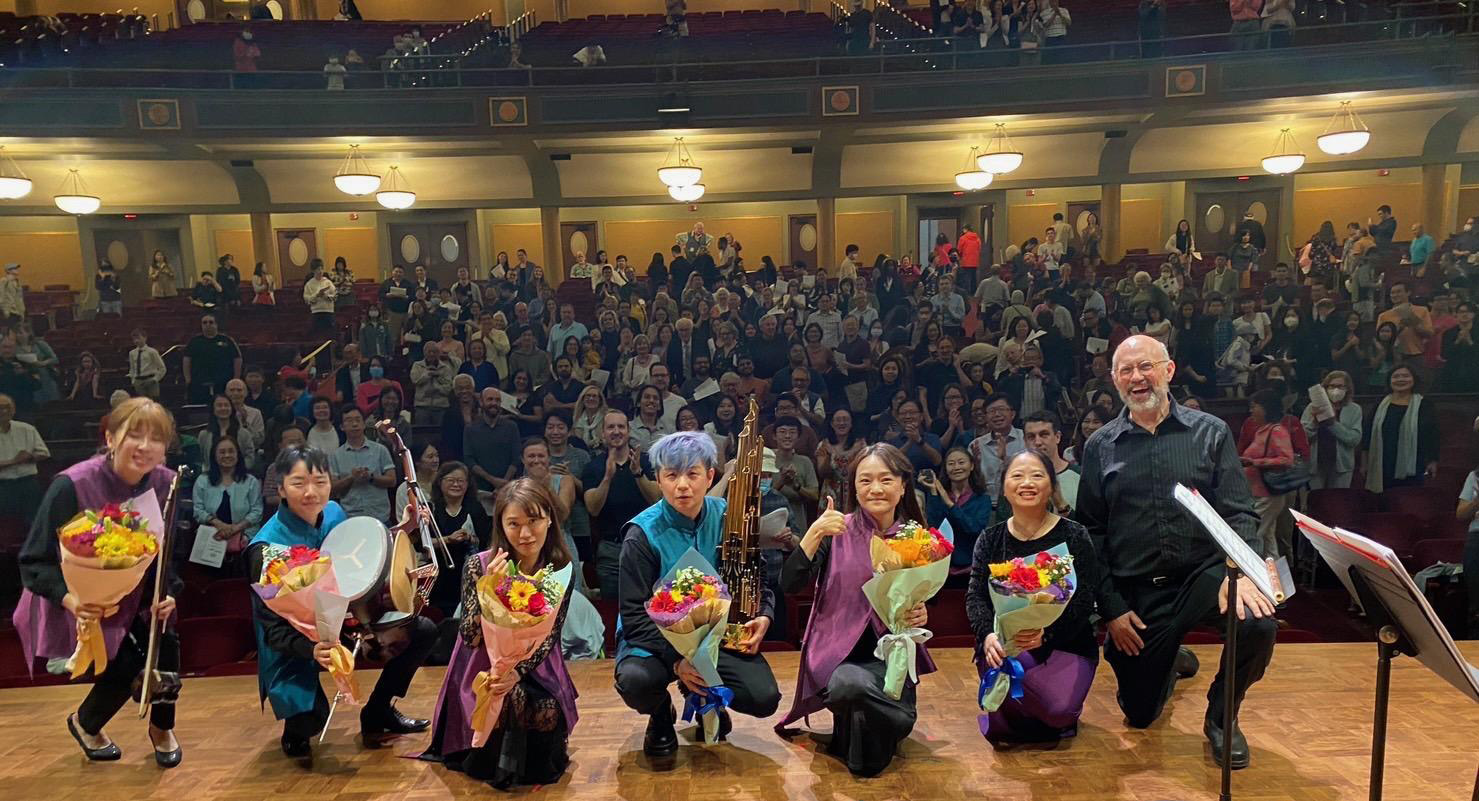 A row of performers holding flowers poses for a bow, with the audience behind them in Hill Auditorium