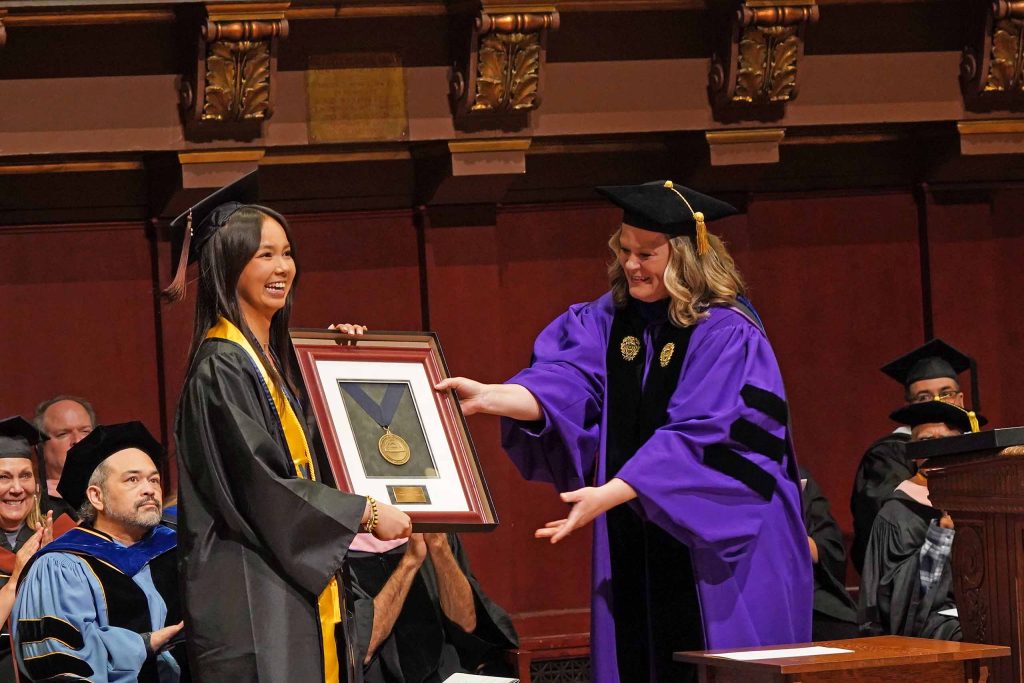 Wearing academic regalia standing on stage at Hill Auditorium, Alexandra Humphreys holds up a framed medal just handed to her by Kate Fitzpatrick