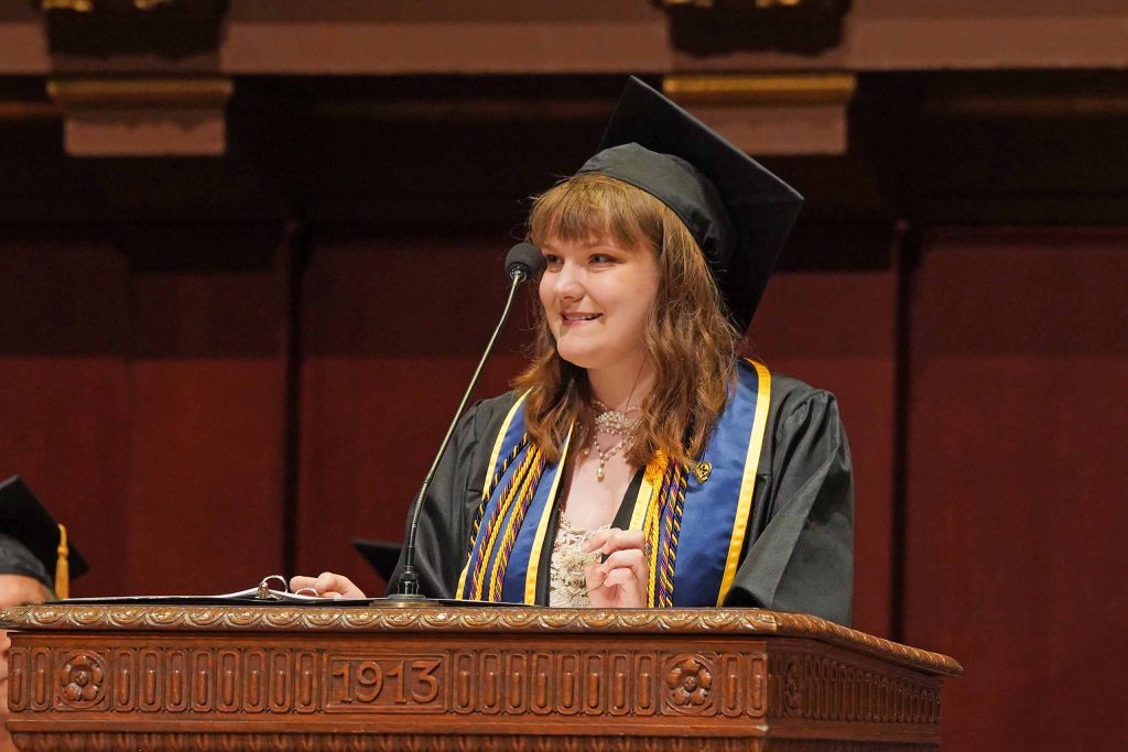 Grace Lutenske speaks from the podium marked 1913 at Hill Auditorium, wearing academic regalia