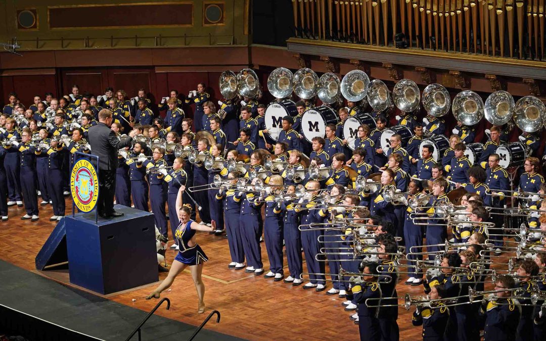 On the stage of Hill Auditorium, the Michigan Marching Band stands in performance. The conductor faces them on a raised block, while a baton twirler performs at the front.