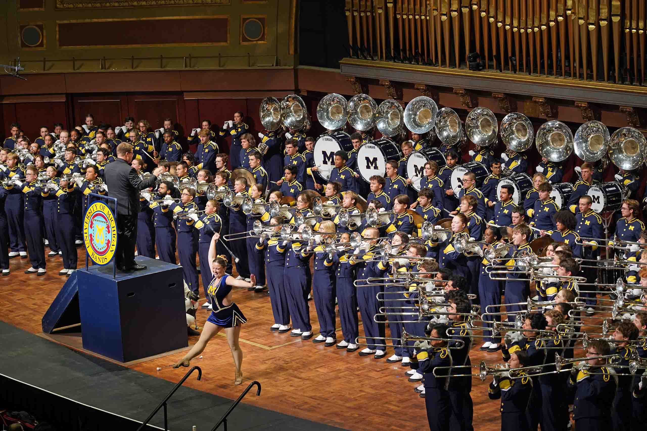 On the stage of Hill Auditorium, the Michigan Marching Band stands in performance. The conductor faces them on a raised block, while a baton twirler performs at the front.