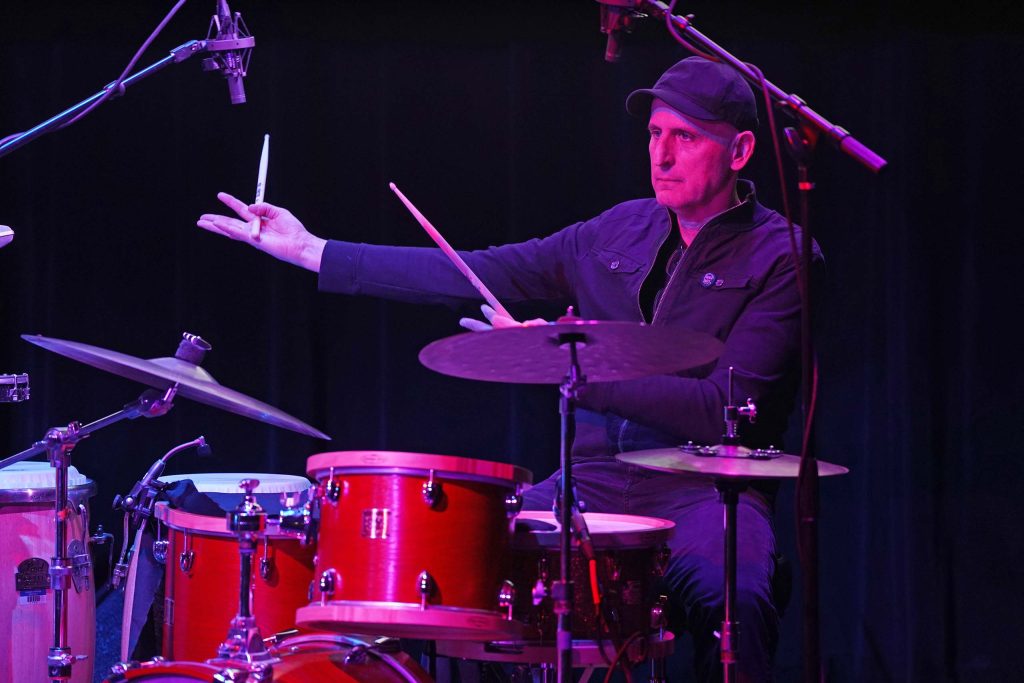 Michael Gould performs seated at a drumset on a darkly lit stage, gesturing to something nearby