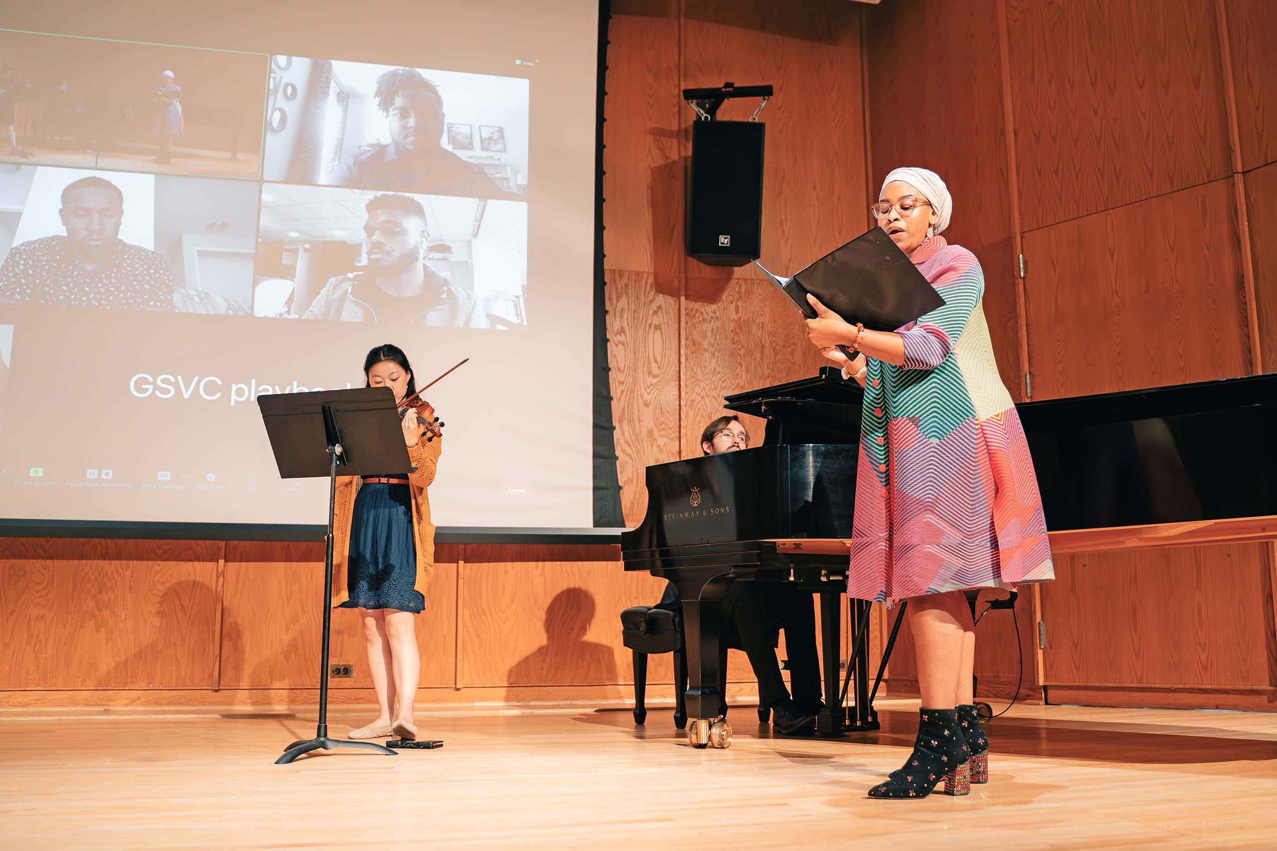 On the stage of Britton Recital Hall, a vocalist, pianist, and string player perform while faces on a Zoom meeting screen are projected behind them.