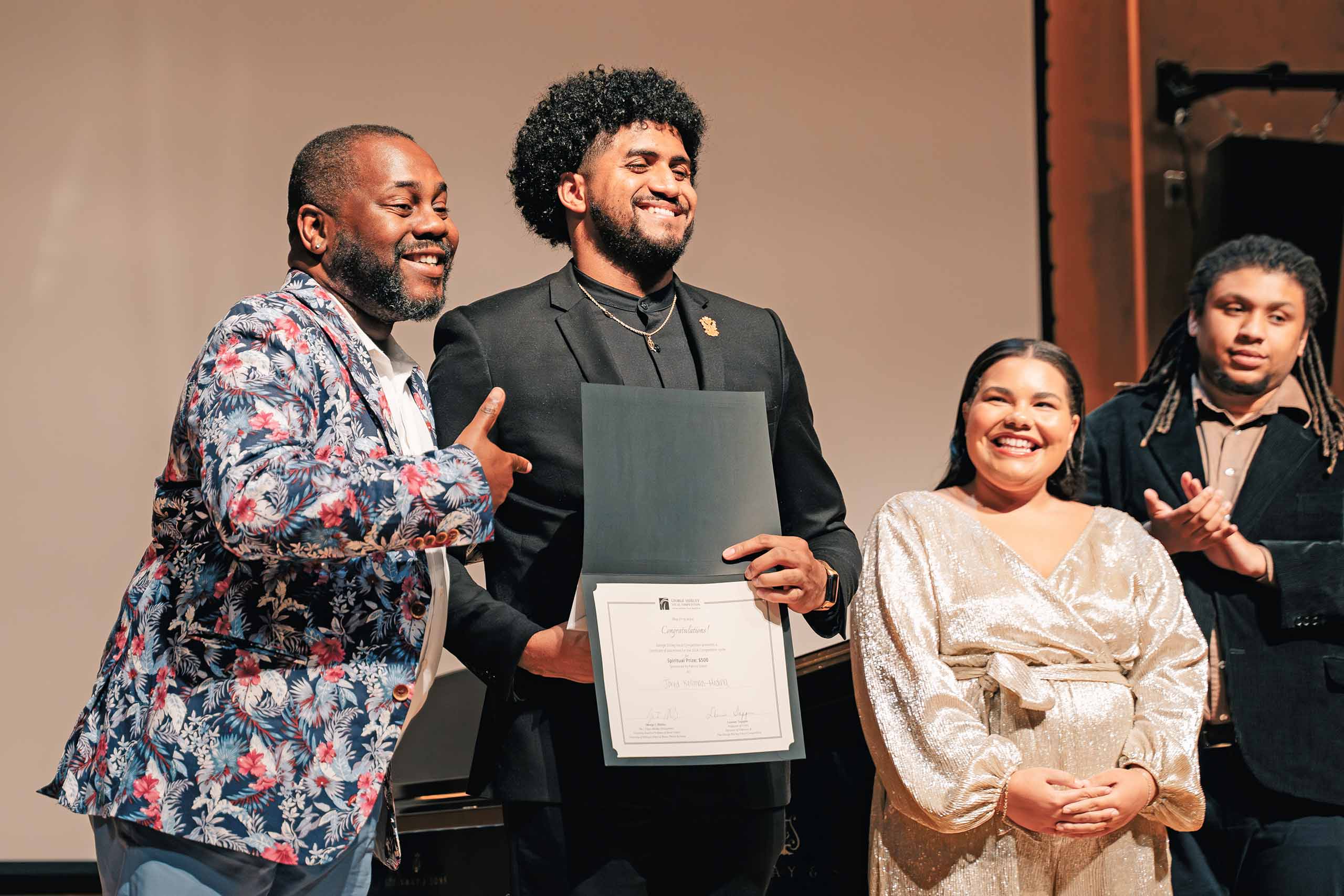 Standing on stage and smiling for a photo, Jared Kellum Medina holds up his GSVC prize certificate while donor Kenneth Overton points to it