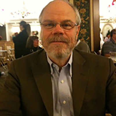 Photo of James Borders seated in a social space, wearing a brown suit coat and collared shirt