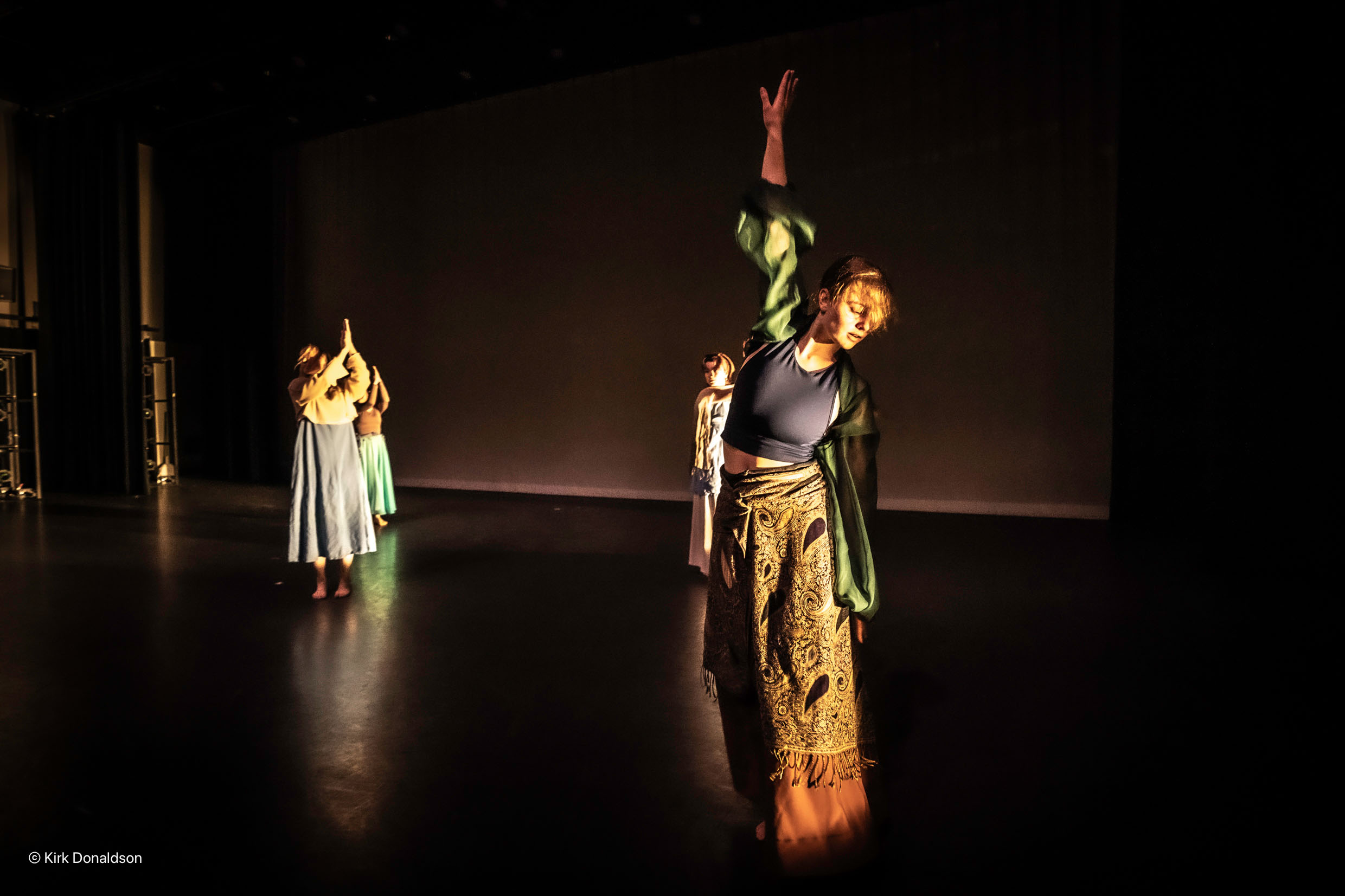 Ladina Schaller reaches an arm high up while performing with other dancers on a dark stage; all are attired in long flowing costumes with neutral tones.