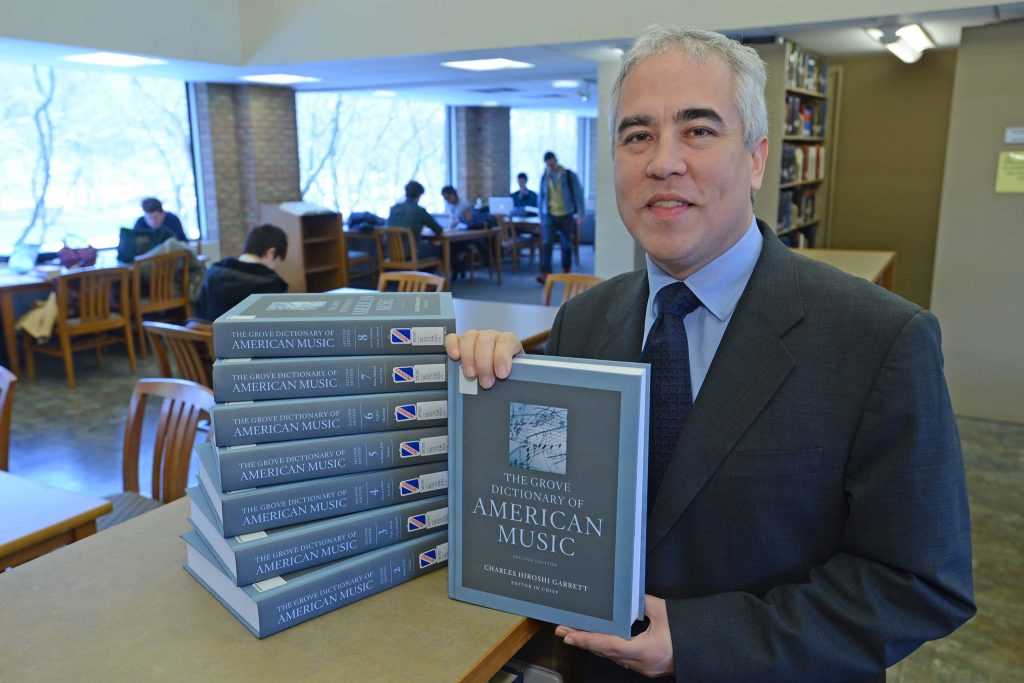 Pictured in the library at SMTD, Charles Hiroshi Garrett holds up a copy of the New Grove Dictionary of American Music, next to a stack of the books on a table.