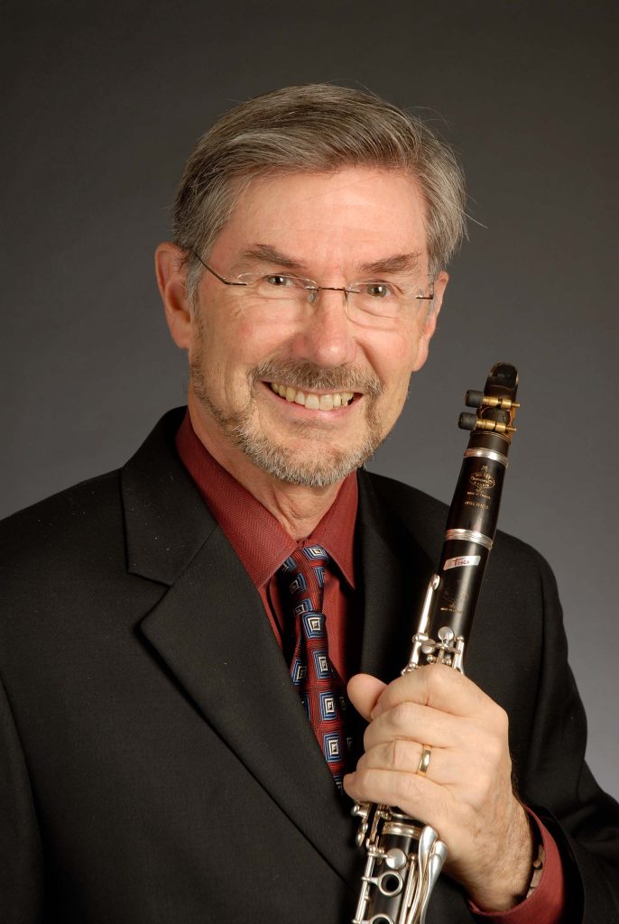 Studio portrait of Fred Ormand holding his clarinet and wearing a black suit coat with red shirt and a tie; grey background.