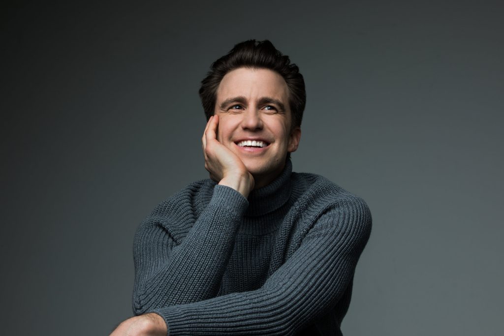 Studio portrait of Gavin Creel resting chin on his hand, wearing grey turtleneck, with a grey backdrop.