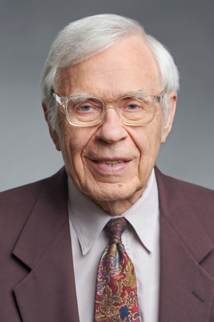 Studio portrait of Paul Lehman wearing a brown suit coat and tie; light grey background.