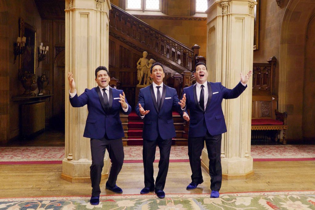 Three singers pose standing with palms raised, wearing matching attire of dark blue suit coats with black lapels and ties; stone columns, a statue, and a carved wooden staircase in the background