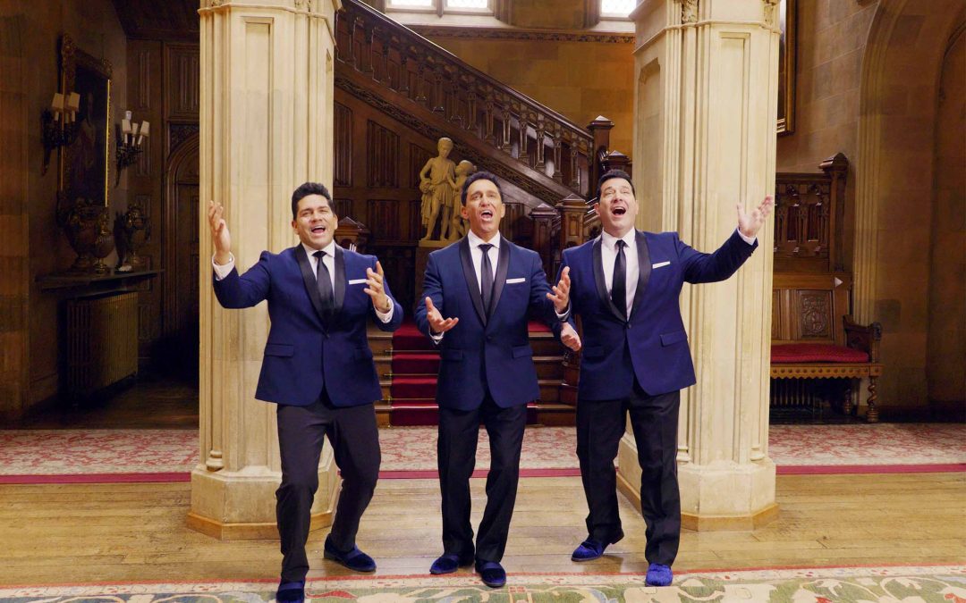 Three singers pose standing with palms raised, wearing matching attire of dark blue suit coats with black lapels and ties; stone columns, a statue, and a carved wooden staircase in the background