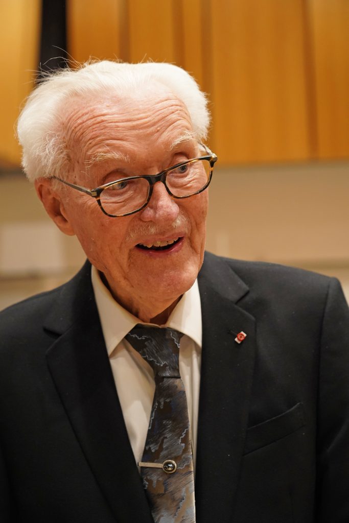 In an action photo taken at SMTD, William Malm wears dark-rim glasses, dark suit coat and tie.