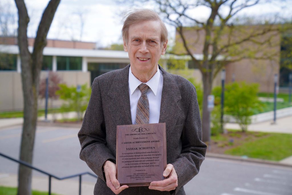 Photo of Yizhak Schotten taken outside facing the Moore Building; Schotten is wearing a brown suit coat and tie and is holding an award plaque.