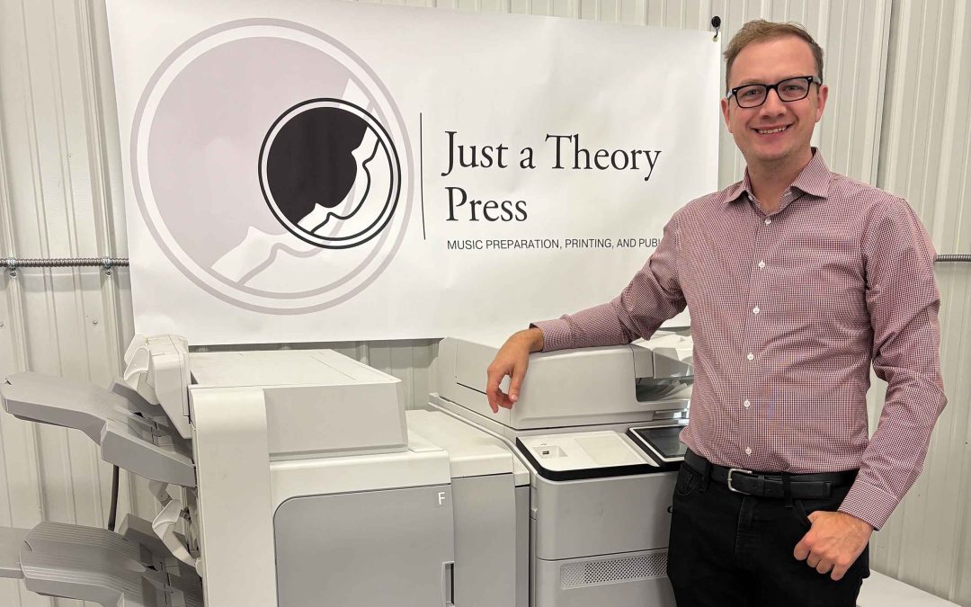 Joshua DeVries poses next to an industrial printer with "Just a Theory Press" banner hanging in backdrop.