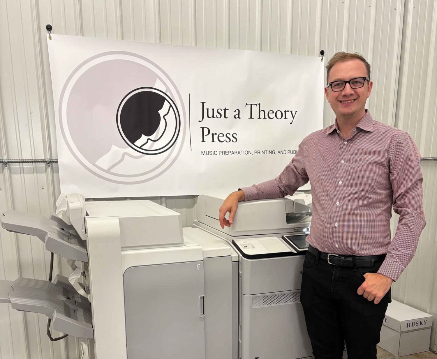 Joshua DeVries poses next to an industrial printer with "Just a Theory Press" banner hanging in backdrop.