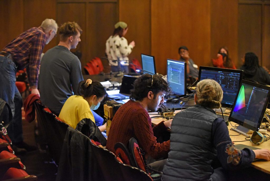 Around 10 people are working and talking with eachother, seated in rows of red theatre seats with a long table, four computer monitors and other equipiment.