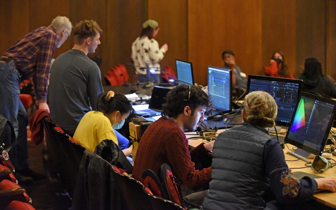 Around 10 people are working and talking with eachother, seated in rows of red theatre seats with a long table, four computer monitors and other equipiment.