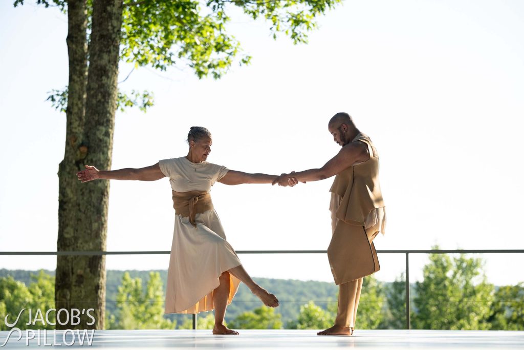 On an outdoor stage with a view of forested hills, two dancers perform with linked arms, costumed in light colored, flowing natural fabrics.