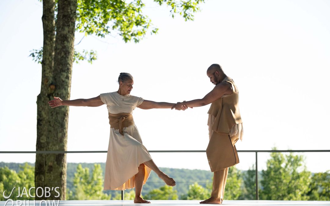 On an outdoor stage with a view of forested hills, two dancers perform with linked arms, costumed in light colored, flowing natural fabrics.