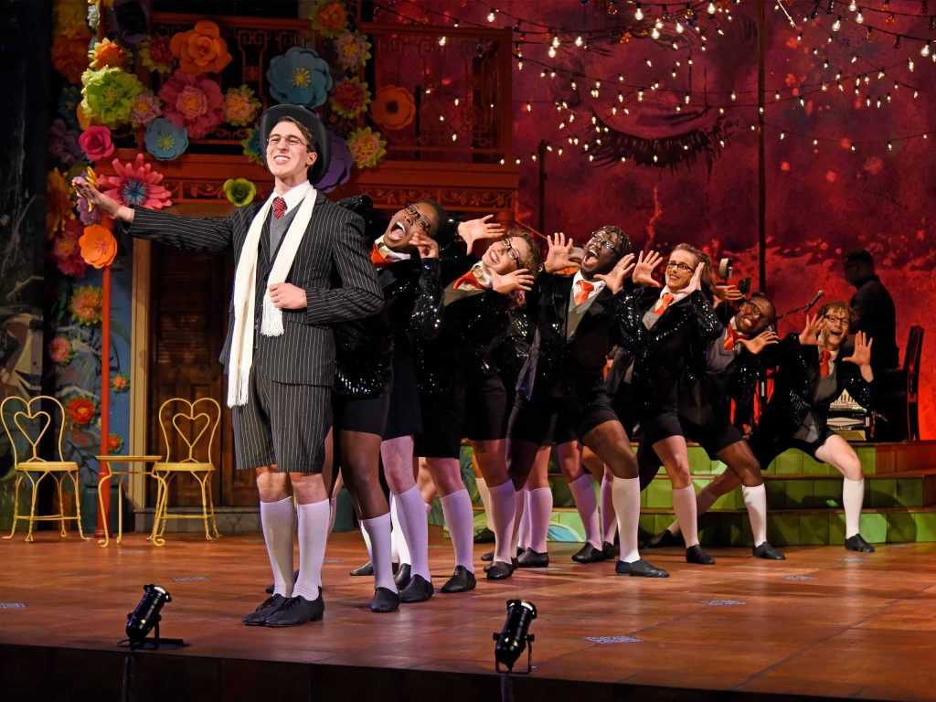 On stage set with colorful flowers and string lights, a cast dressed in school uniforms performs standing in a line, leaning out with jazz hands