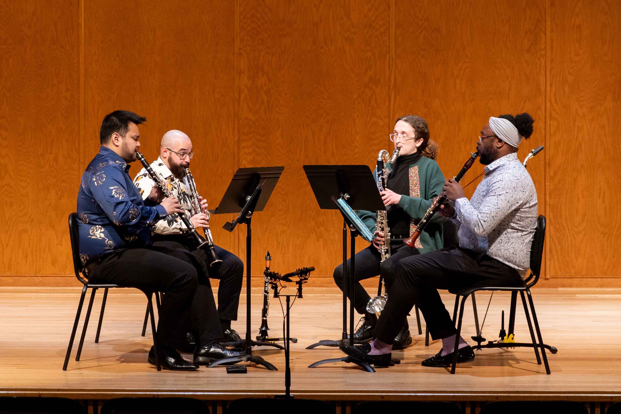 A quartet of two clarinets, bass clarinet, and basset horn performs seated on stage; all dressed in concert attire with floral patterned accents.