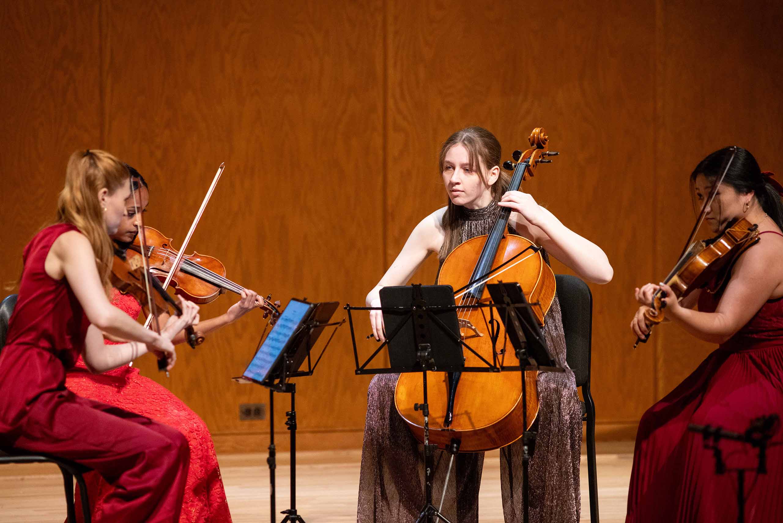 A quartet of 2 violins, cello and viola performs seated on stage, dressed in concert attire including red and pink accents.