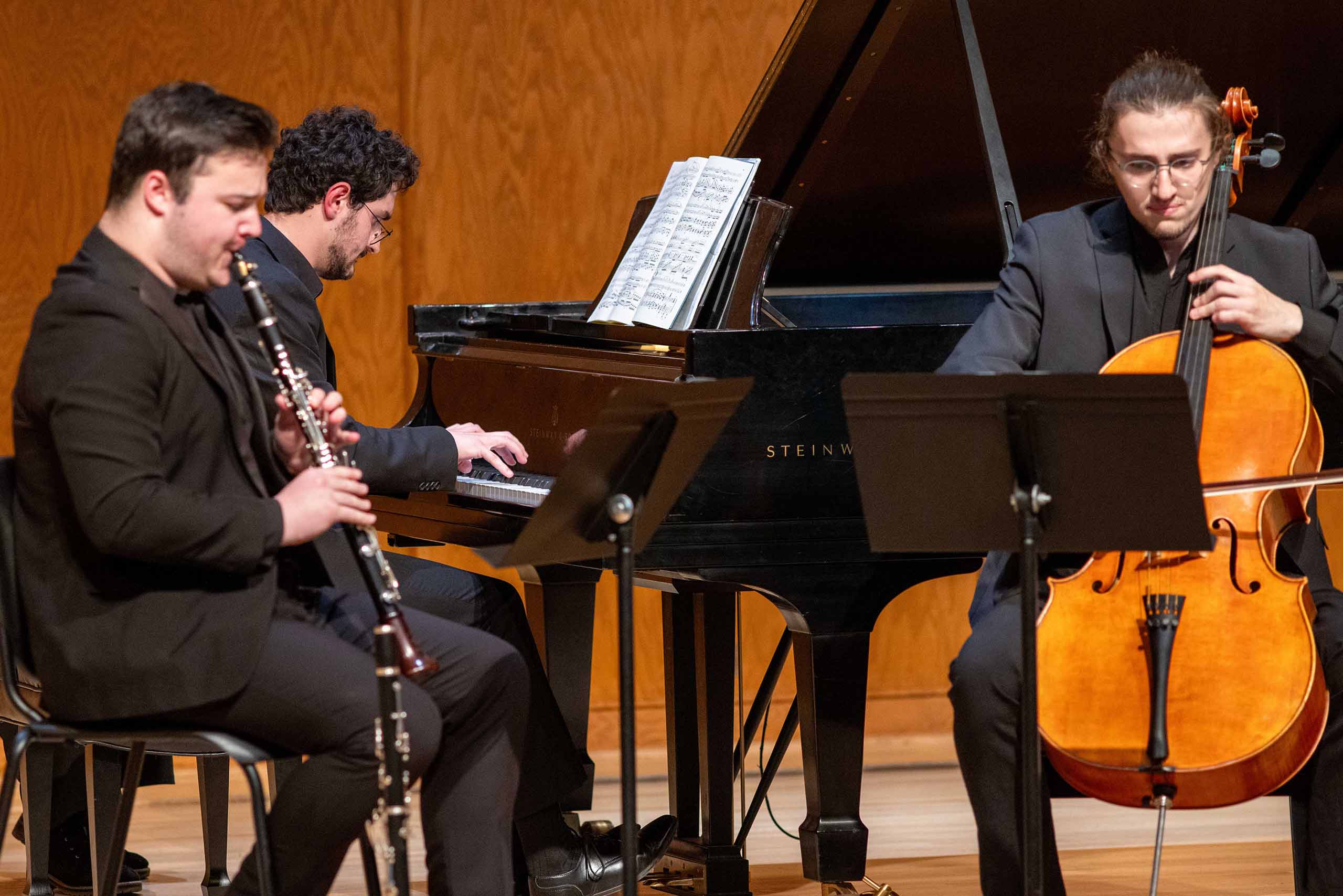 A trio of clarinet, piano, and cello performs seated on stage, dressed in black concert attire