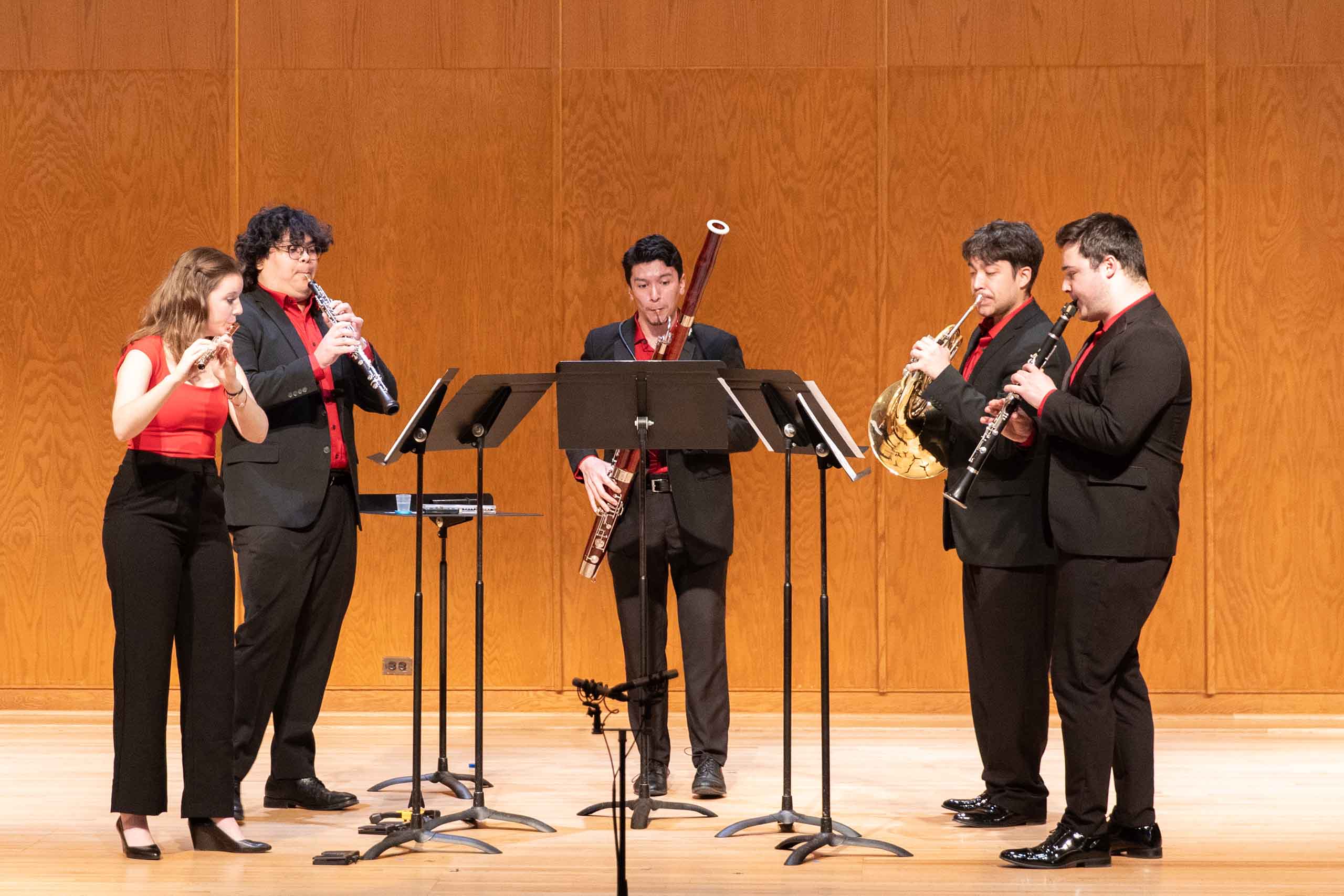 A quintet of flute, oboe, bassoon, French horn, and clarinet perform standing on stage, dressed in concert attire including red accents.