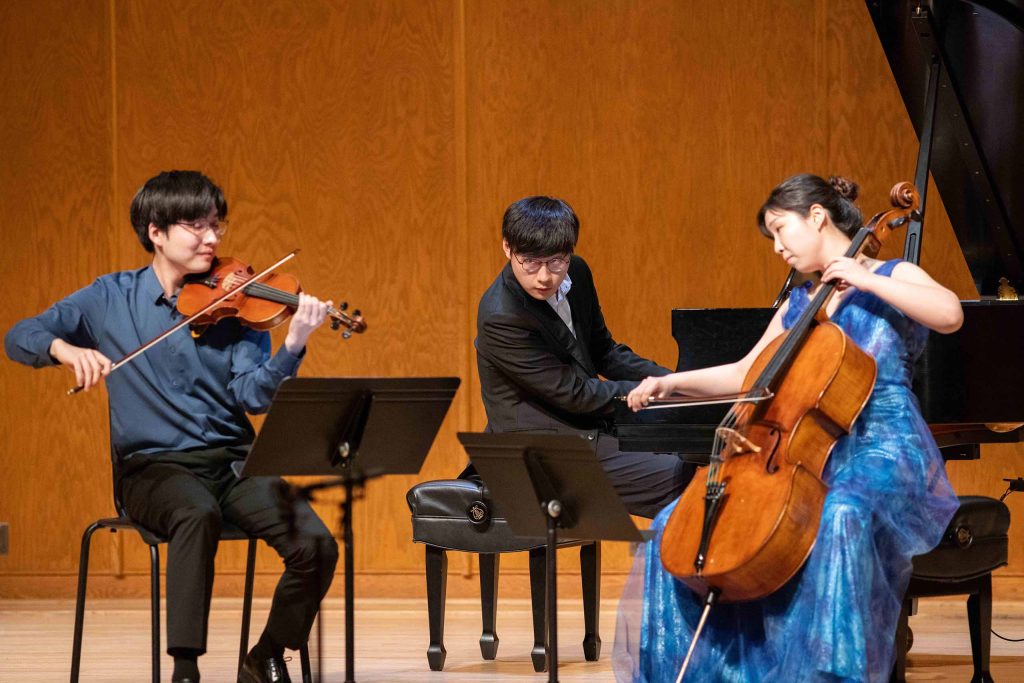 A trio of violin, piano, and cello performs seated on stage, dressed in concert attire including blue accents.