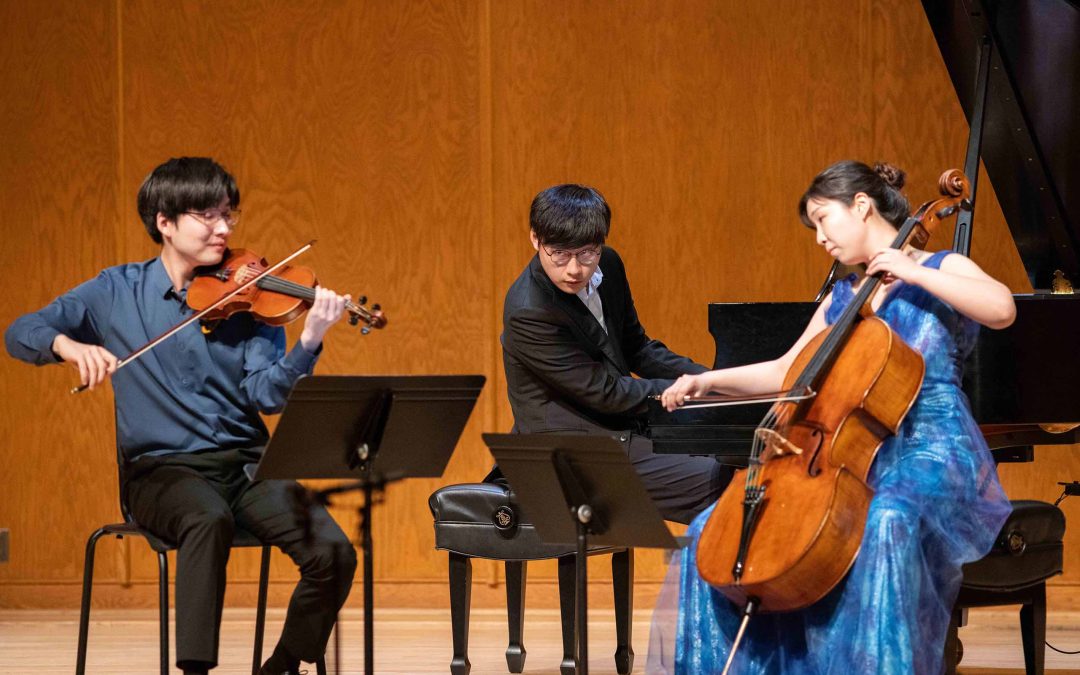 A trio of violin, piano, and cello performs seated on stage, dressed in concert attire including blue accents.