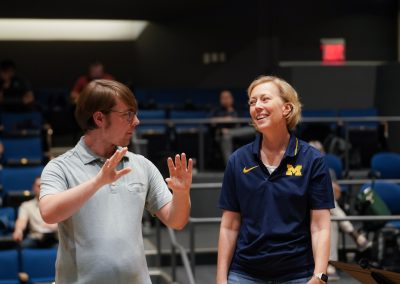 Wearing a Michigan collared shirt, Professor Courtney Snyder stands in conversation with a participant conductor at the 2024 Wind Conducting Workshop, with rows of tiered seating behind them.