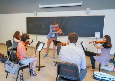 Four adult students seated in a classroom, facing a standing instructor, all playing violins.