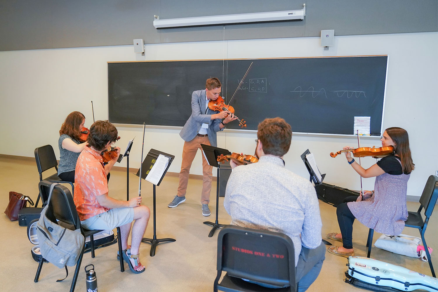 Four adult students seated in a classroom, facing an instructor, all playing violins 