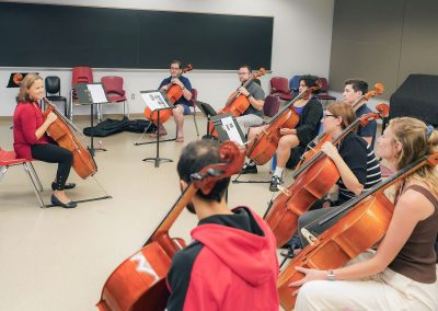 Seven adult students seated in a classroom, facing a seated instructor, all holding cellos.