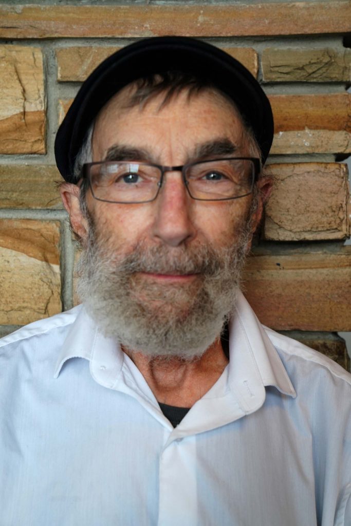 Portrait of Paul Schoenfeld next to a brick wall; wearing a black cap, glasses, and white Oxford shirt.